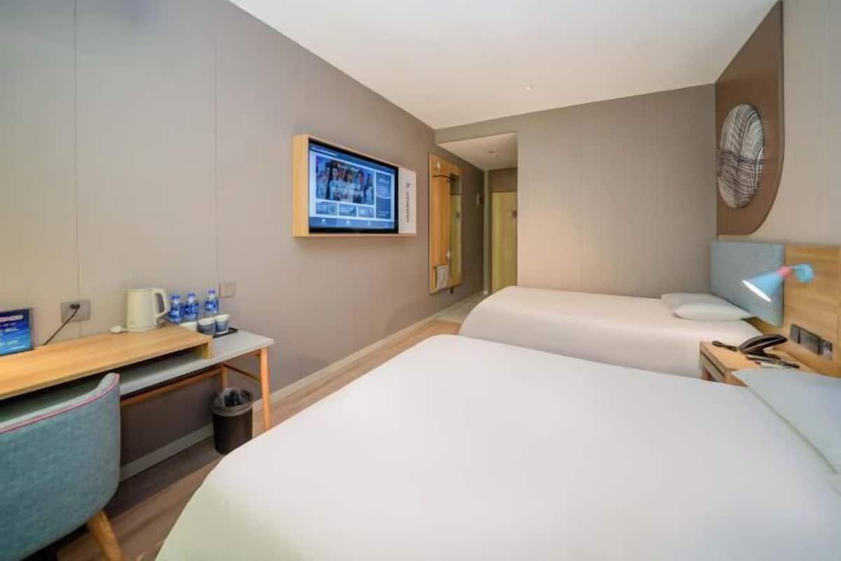 A hotel room with two beds, a desk with a computer, a TV mounted on the wall, and a small trash bin.