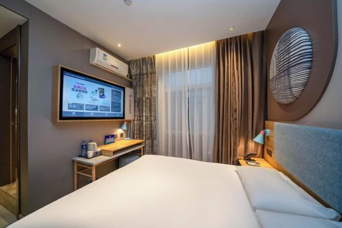 A hotel room with a bed, a desk with a computer, a TV mounted on the wall, and a window with curtains.