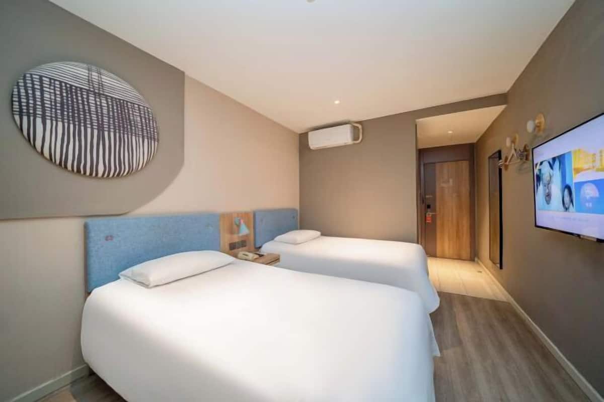 A hotel room with two beds, a TV, a wall-mounted artwork, and a hallway leading to an elevator.