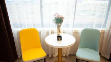 Dos sillas con una pequeña mesa redonda entre ellas, un jarrón con flores sobre la mesa y una ventana con cortinas al fondo.