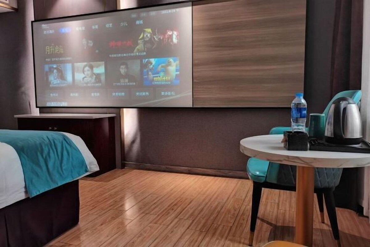 A hotel room with a bed, a TV mounted on the wall displaying movie options, a small round table with a kettle and a water bottle.