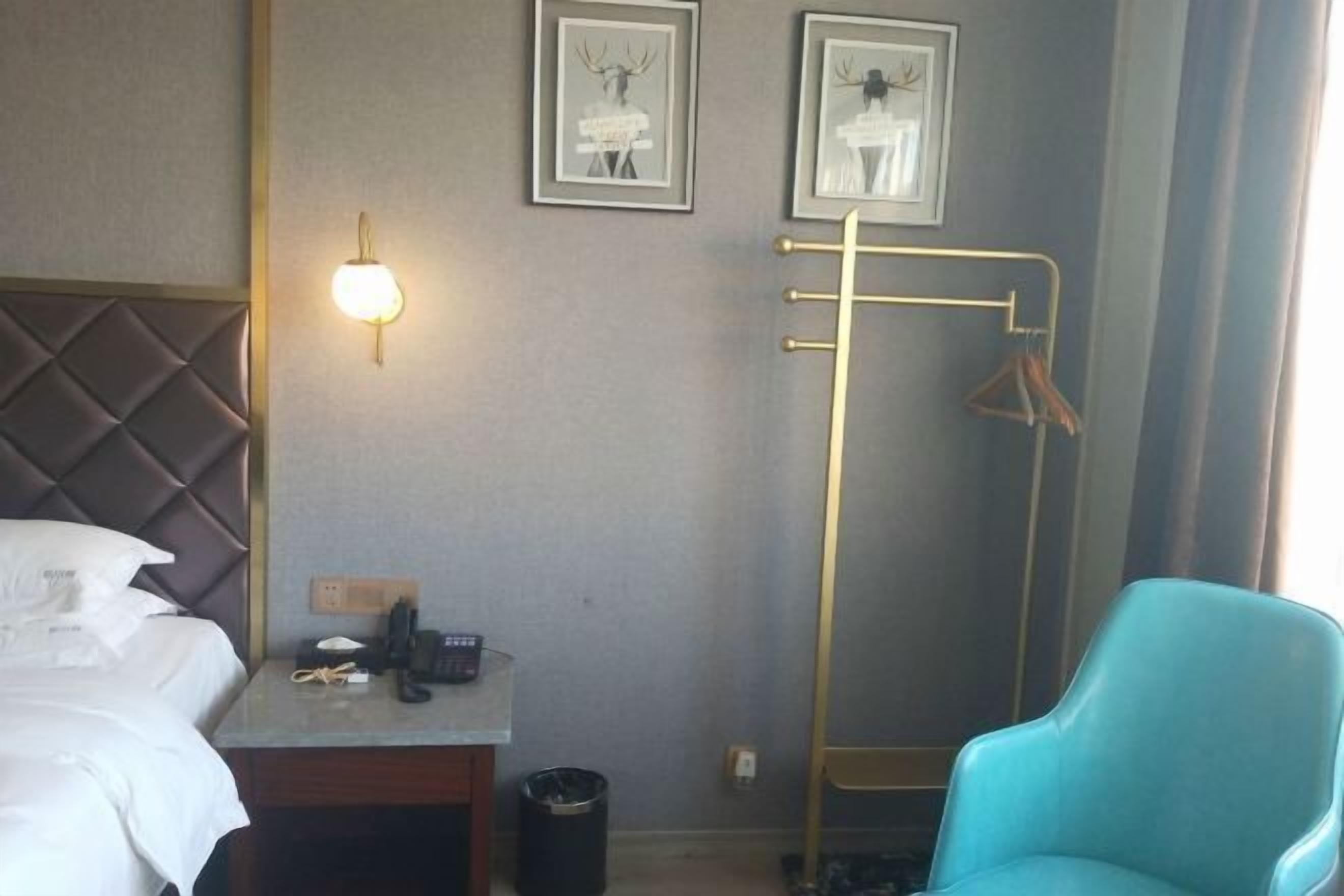 A hotel room with a bed, a desk with a phone, a trash can, a blue chair, and a coat rack with hangers.