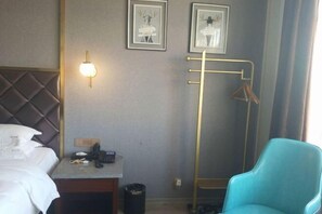 A hotel room with a bed, a desk with a phone, a trash can, a blue chair, and a coat rack with hangers. - Hillman Light Luxury Hotel (Yueyang Taiyangqiao Branch) (Yueyang)