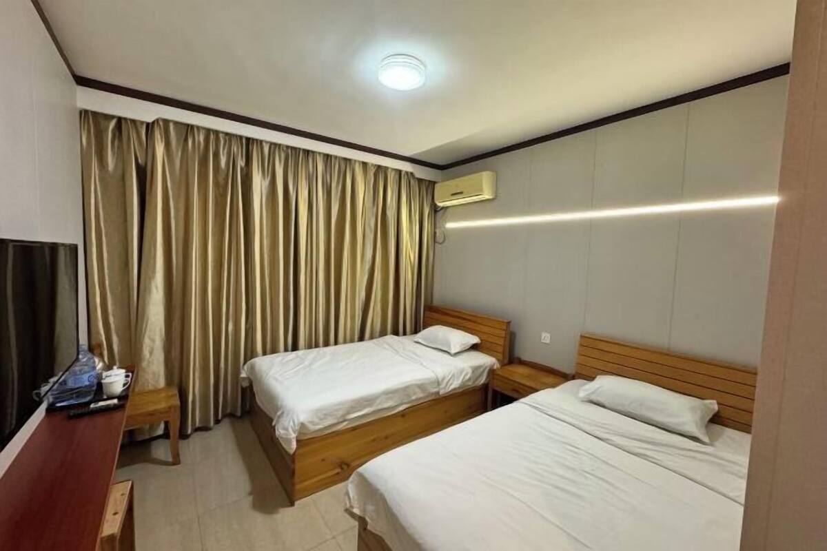 A hotel room with two single beds, a wooden nightstand, a desk with a chair, a window with curtains, and an air conditioning unit.