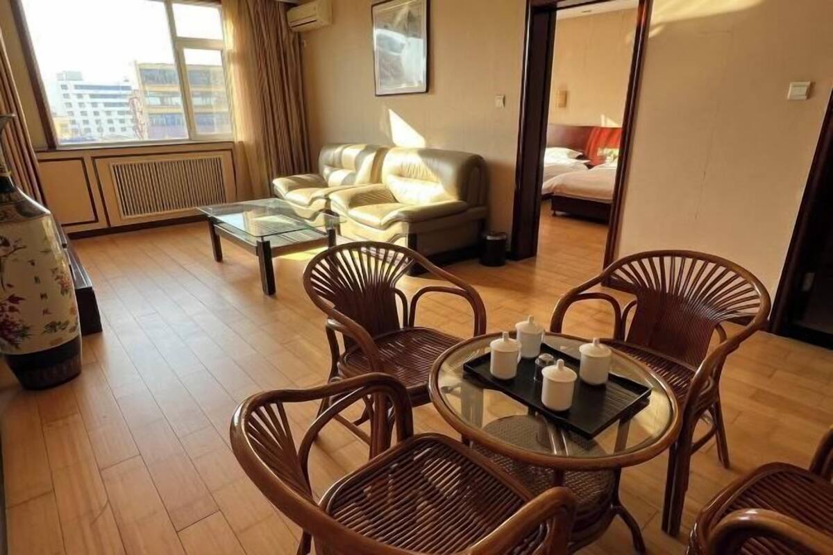 A hotel room with a sofa, a glass coffee table, wicker chairs, a bed, and a view of the city.