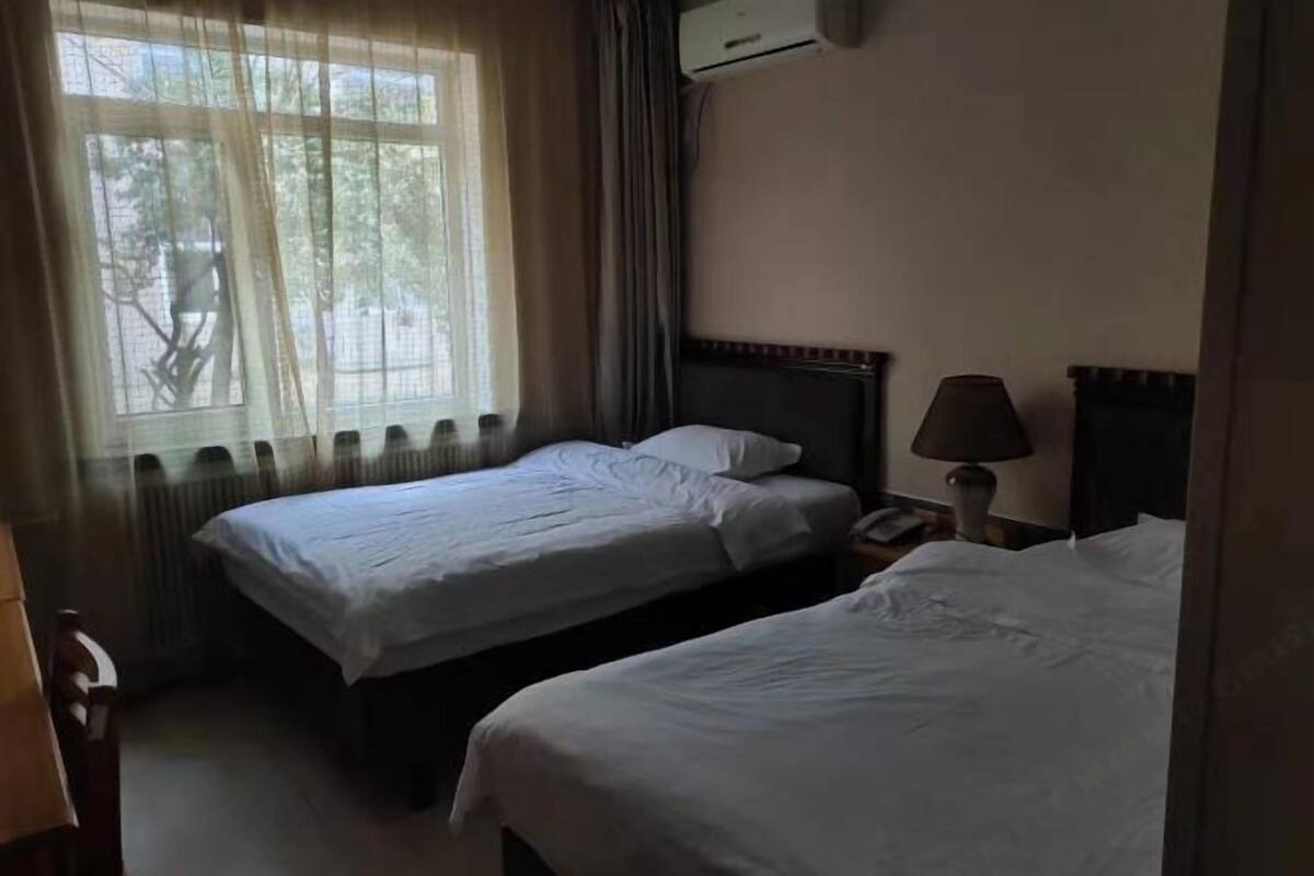 A hotel room with two beds, a window with sheer curtains, an air conditioning unit, and a bedside lamp.