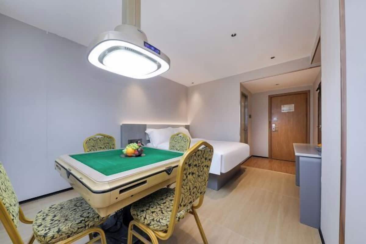 A hotel room with a bed, a desk, a chair, a table with a green tablecloth, and a ceiling light.