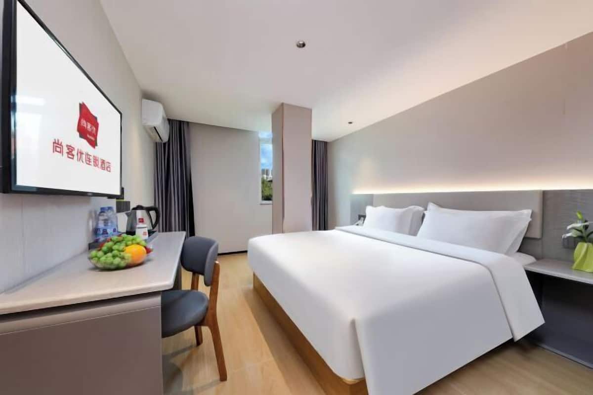 A modern hotel room with a bed, a desk with a chair, a small table with fruits, and a wall-mounted TV.