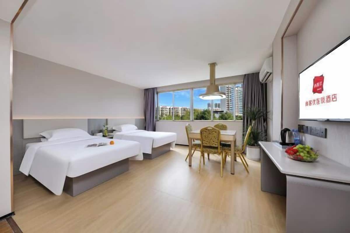 A hotel room with two single beds, a dining table with chairs, a small kitchen area, and a large window offering a city view.