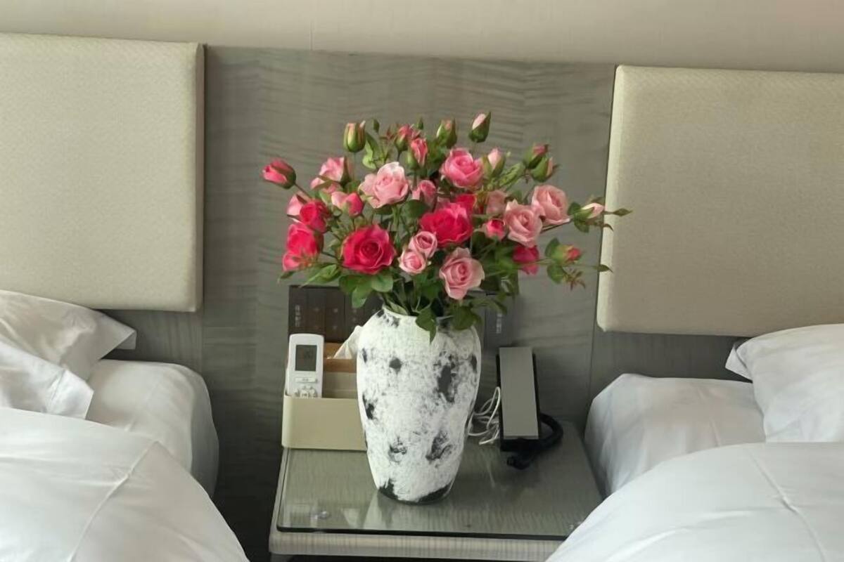 A hotel room with a bed, a bedside table, a vase with pink roses, a phone, and a small electronic device.
