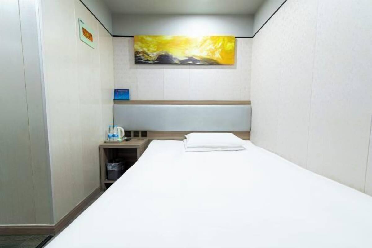 Une chambre d’hôtel avec un lit, une table de chevet, un tableau et une petite étagère sur laquelle se trouvent une bouteille et une boîte.