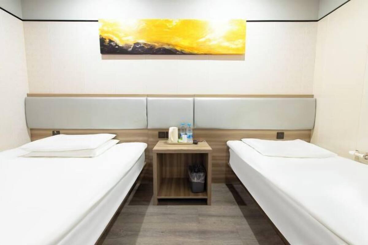 Une chambre d’hôtel avec deux lits simples, une table de chevet en bois avec une poubelle, une petite table avec des articles de toilette et un tableau au mur.