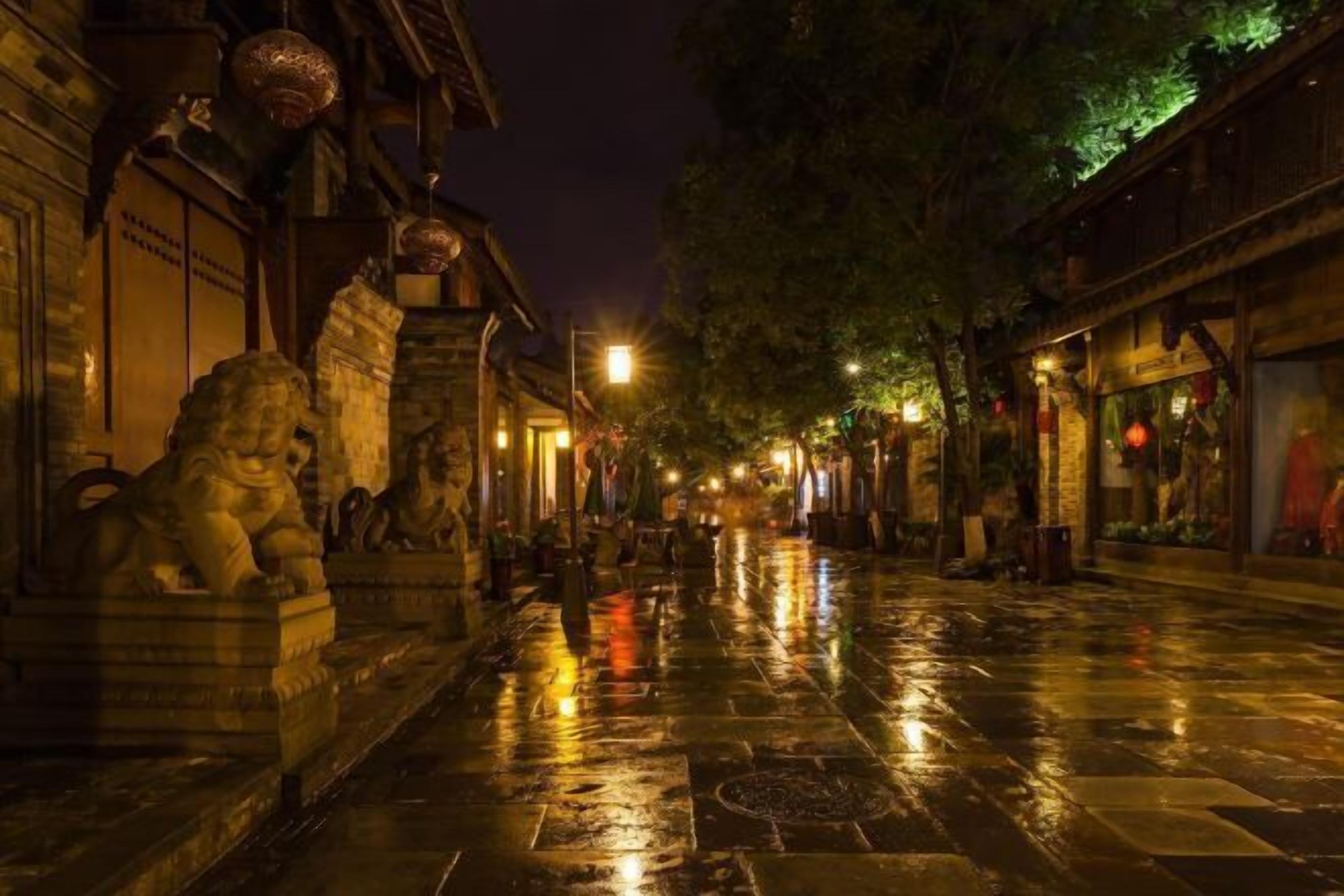 Eine nass glänzende Straße bei Nacht, gesäumt von traditioneller Architektur und steinernen Löwenstatuen.