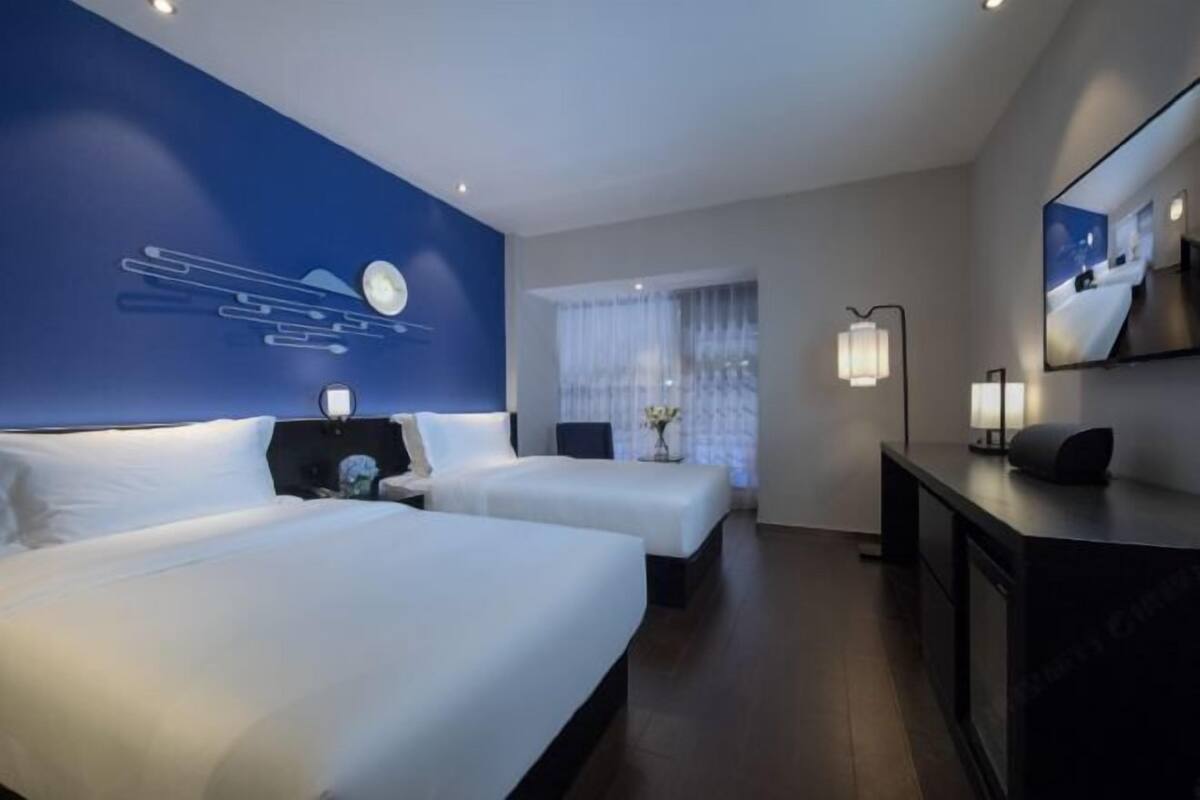 A hotel room with two beds, a desk, a mirror, and a large window with curtains.