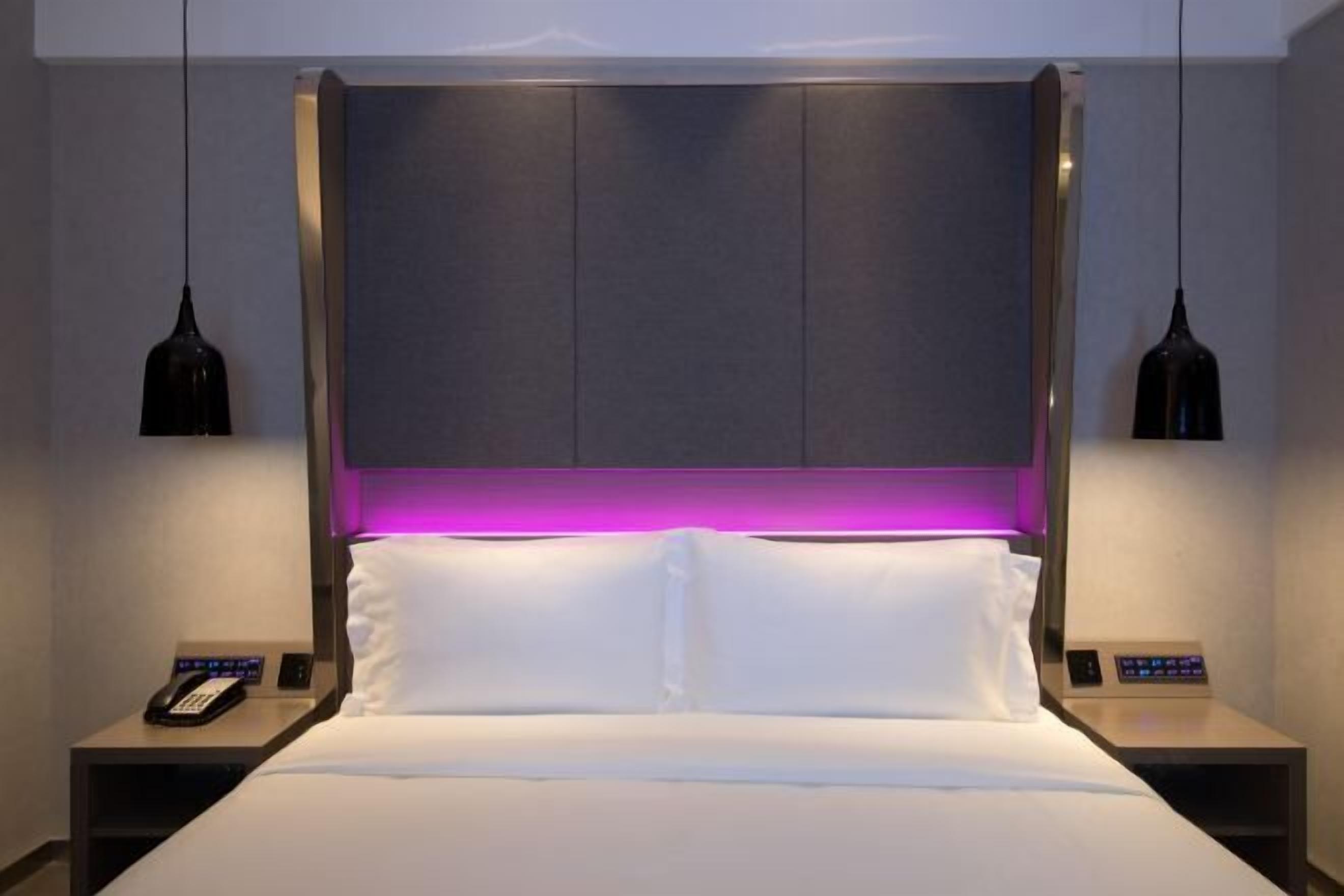 A modern hotel room with a large bed, a headboard with a purple backlight, bedside tables with phones, and pendant lights.