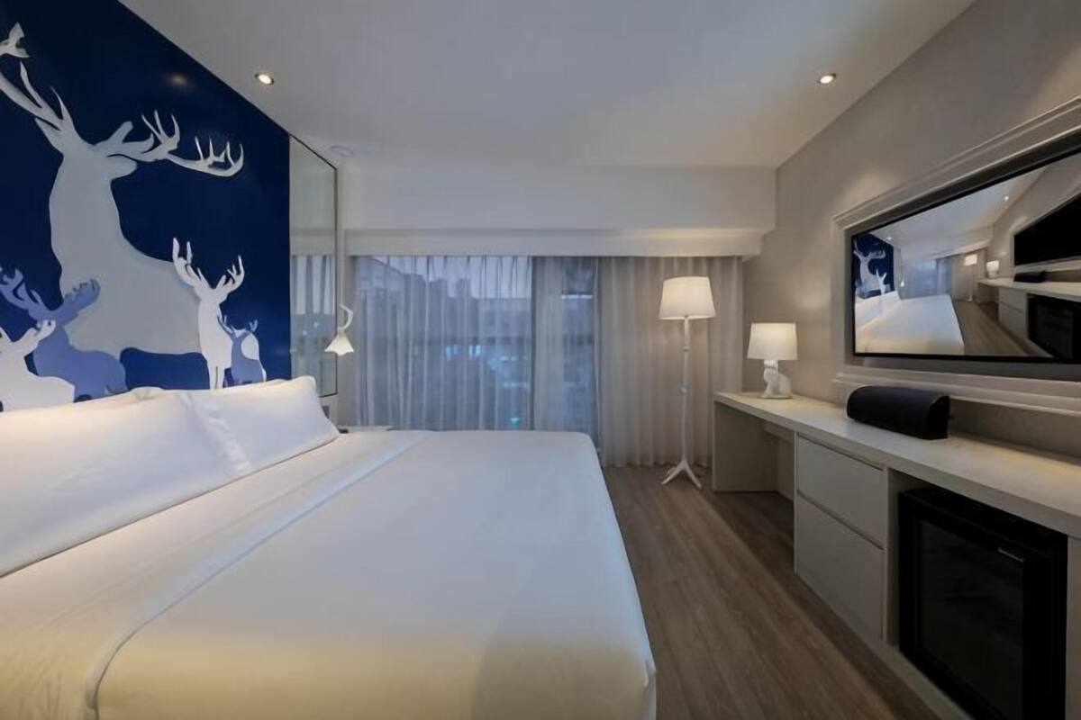 A hotel room with a large bed, a flat-screen TV, a desk with a lamp, and a view of the city through large windows.