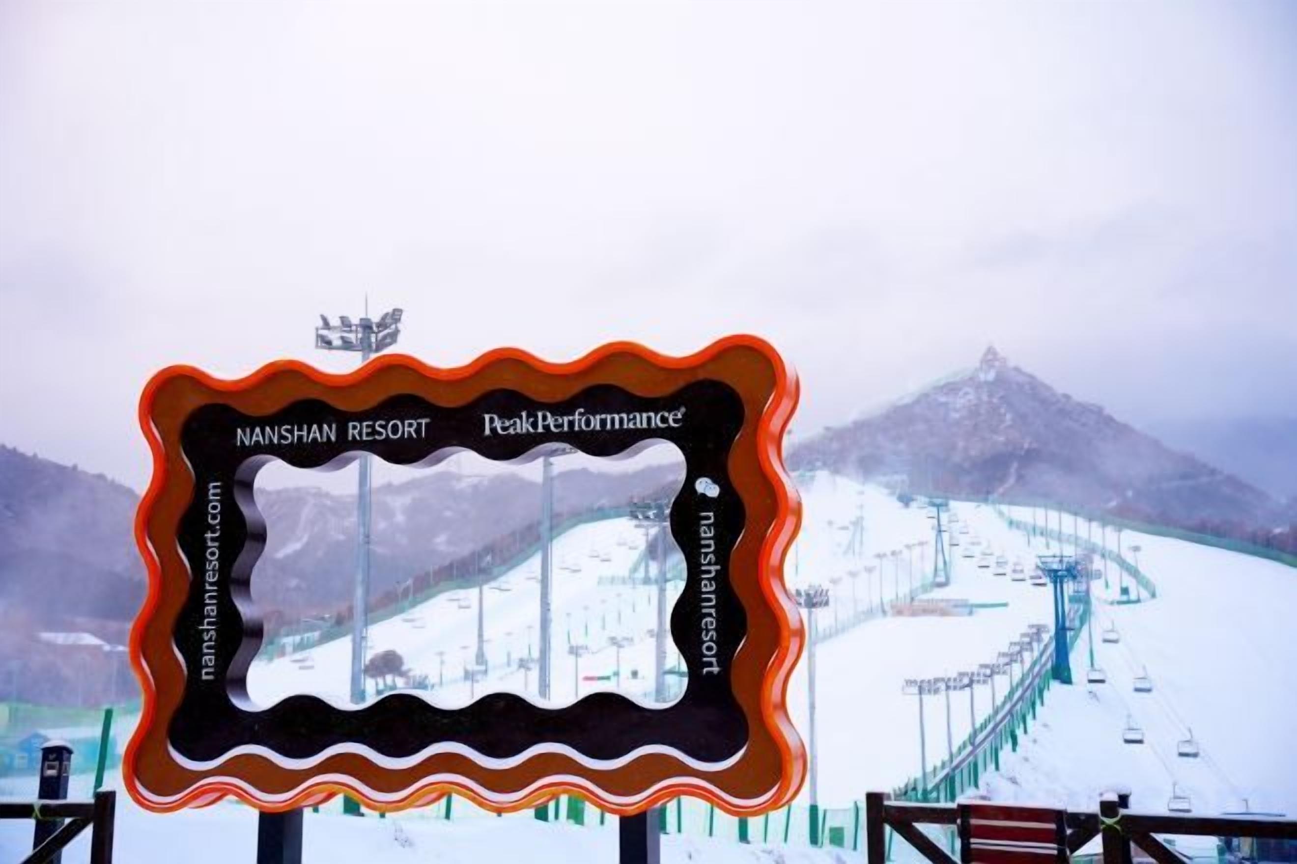 Une enseigne avec les inscriptions « Nanshan Resort » et « PeakPerformance », un paysage de montagne enneigé en arrière-plan et un télésiège.