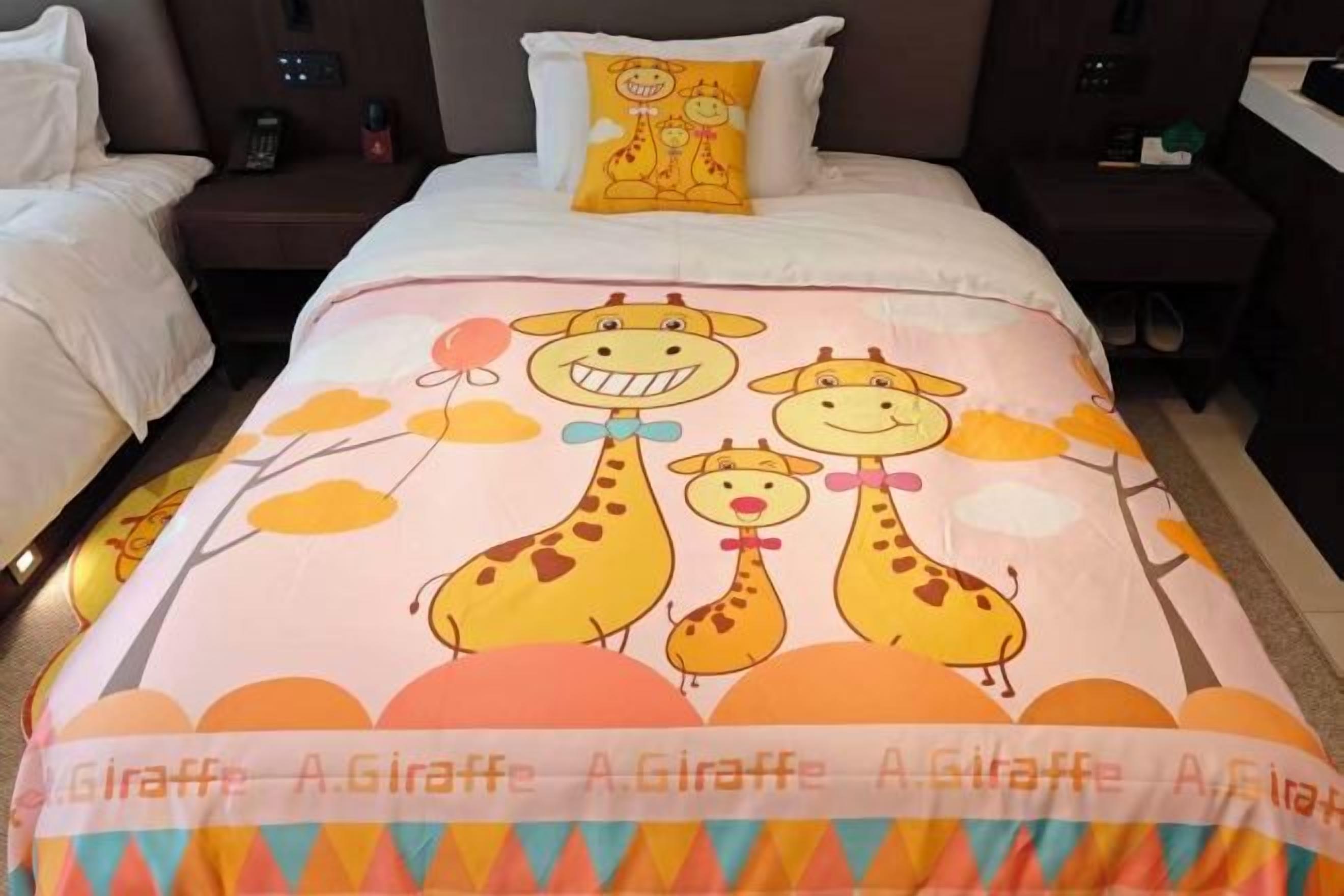 A hotel room with a bed featuring a giraffe-themed bedspread, a nightstand with a lamp, and a bedside table with a phone.