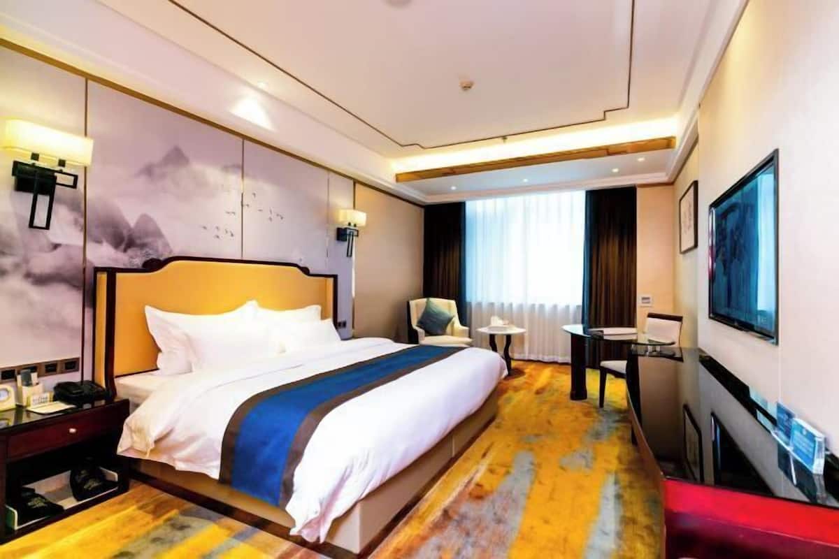 A hotel room with a large bed, a desk, a chair, a TV, and a wall with a mountain painting.