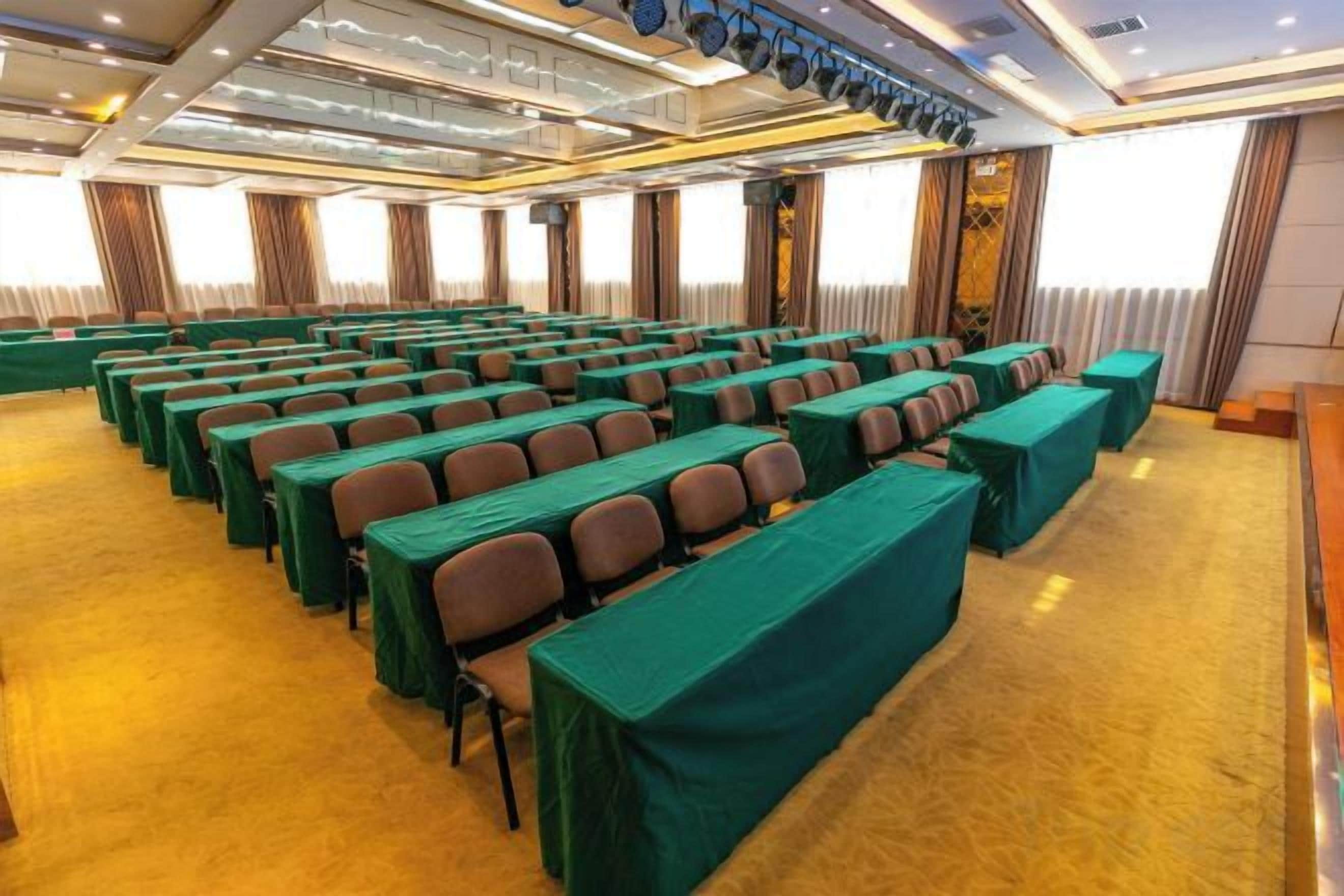 Un salón de conferencias con filas de mesas y sillas, piso alfombrado y un ventanal con cortinas.