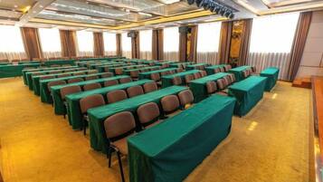 Un salón de conferencias con filas de mesas y sillas, piso alfombrado y un ventanal con cortinas.