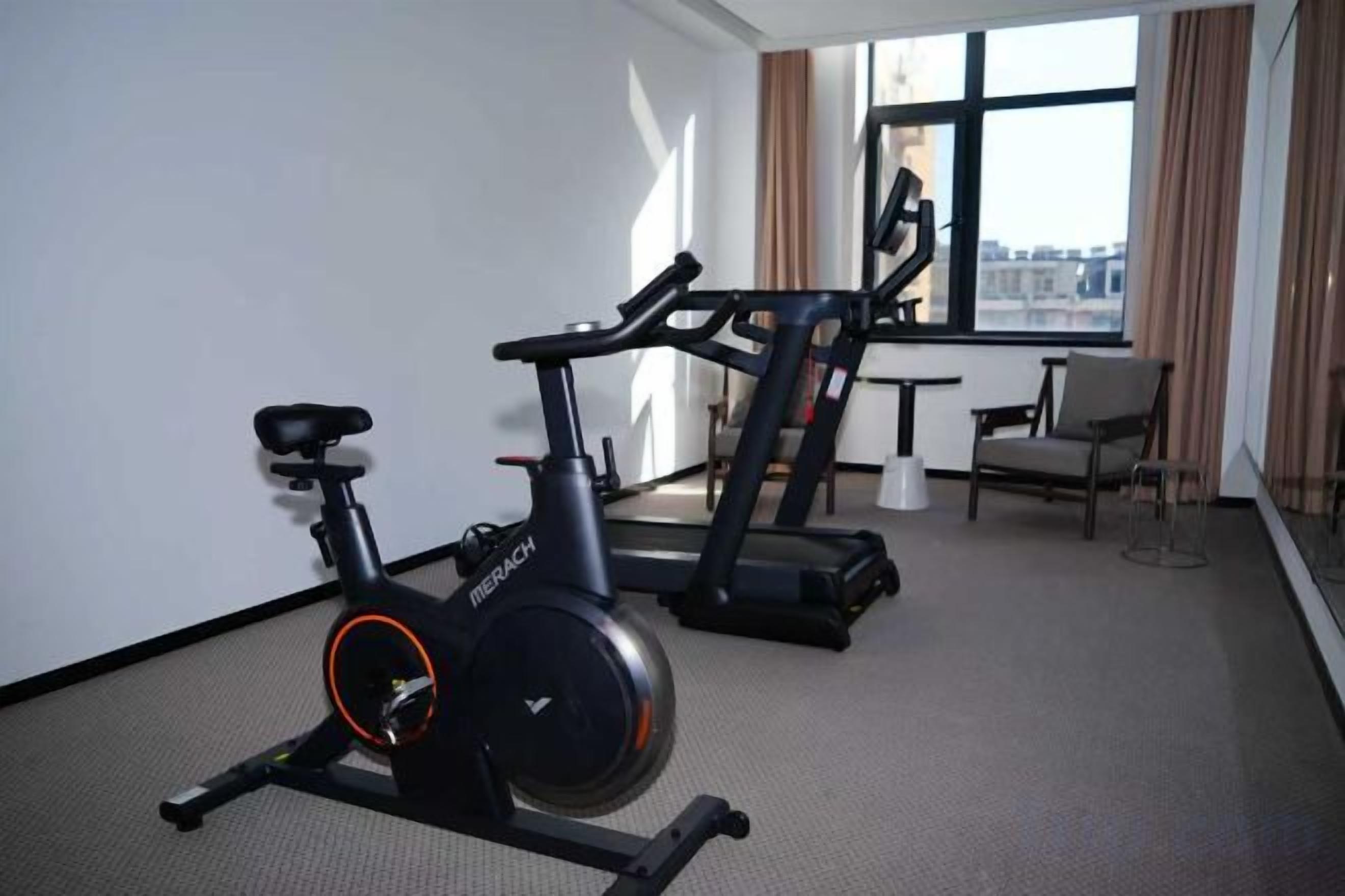A hotel room with a stationary bike, a treadmill, and a view of the city through large windows.