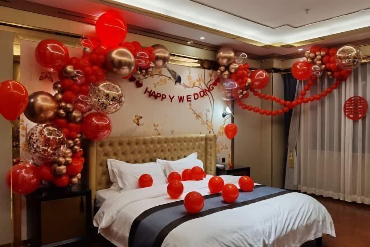 A hotel room with a bed, a nightstand, and a wall decorated with red and gold balloons and a 'Happy Wedding' sign.