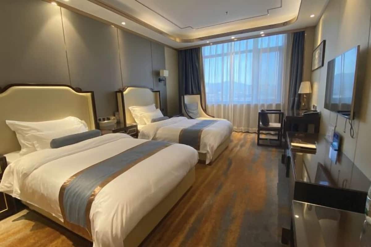 A hotel room with two beds, a desk, a chair, a TV, and a large window.