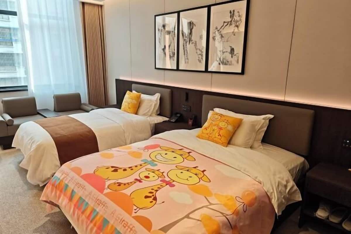 A hotel room with two beds, a large window with curtains, a wall with framed artwork, and a bedside table with shoes.