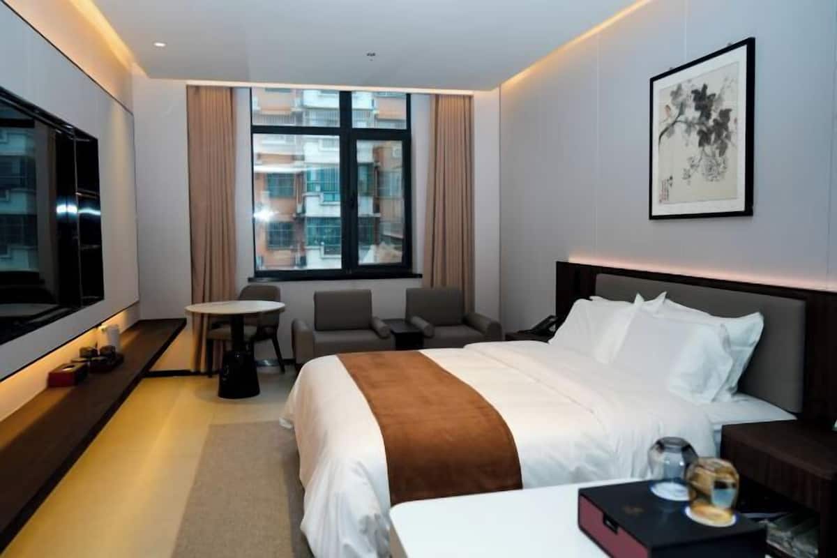 A hotel room with a large bed, a desk, a chair, a TV, and a window with a view of buildings.