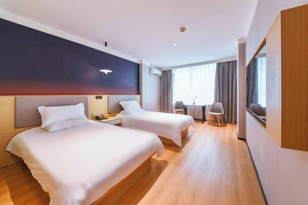 A hotel room with two beds, a desk, a chair, and a large window with curtains.
