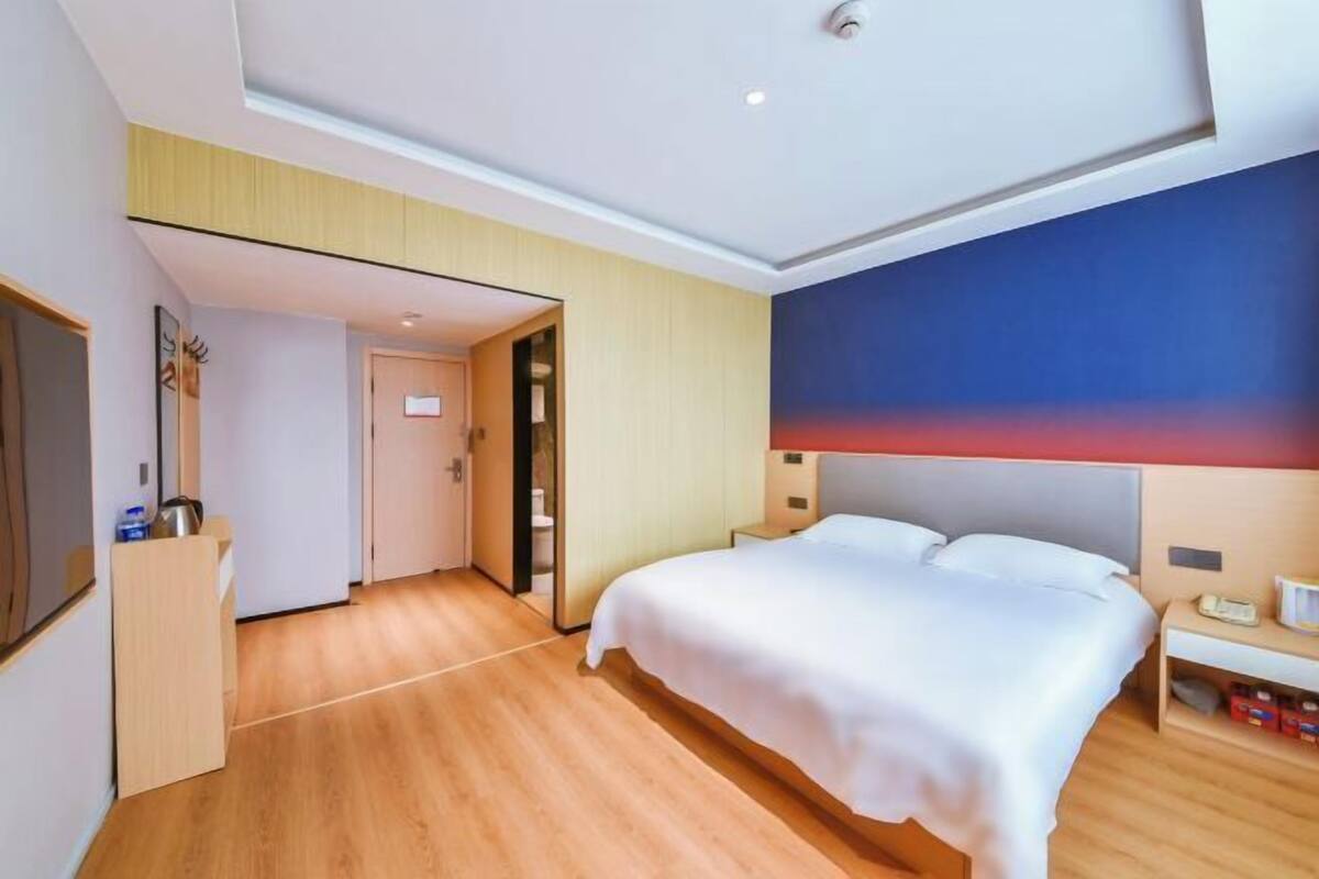 A modern hotel room with a large bed, wooden flooring, a door leading to a bathroom, and a wall with a gradient design.
