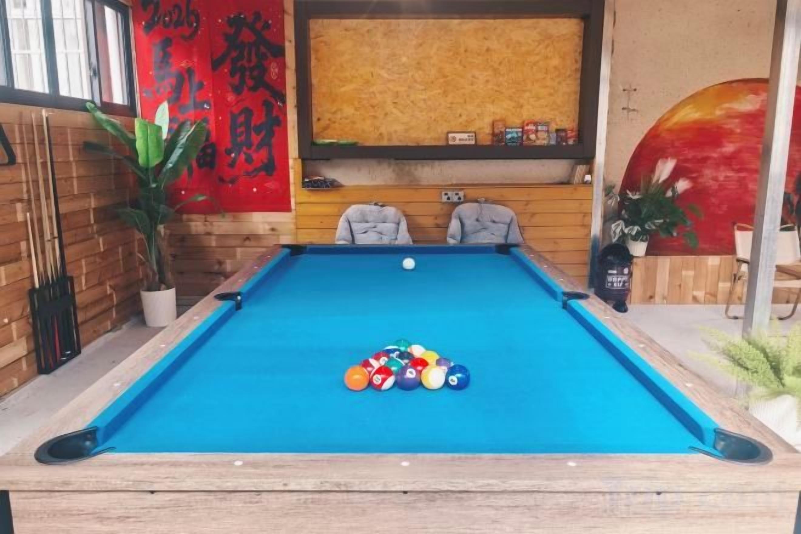 A pool table with balls, two chairs, a red wall with Chinese characters, and a wooden wall with shelves.