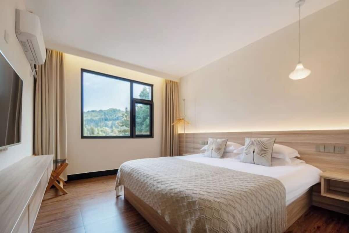 A hotel room with a large bed, a bedside table, a nightstand, a window with a view, and an air conditioning unit.