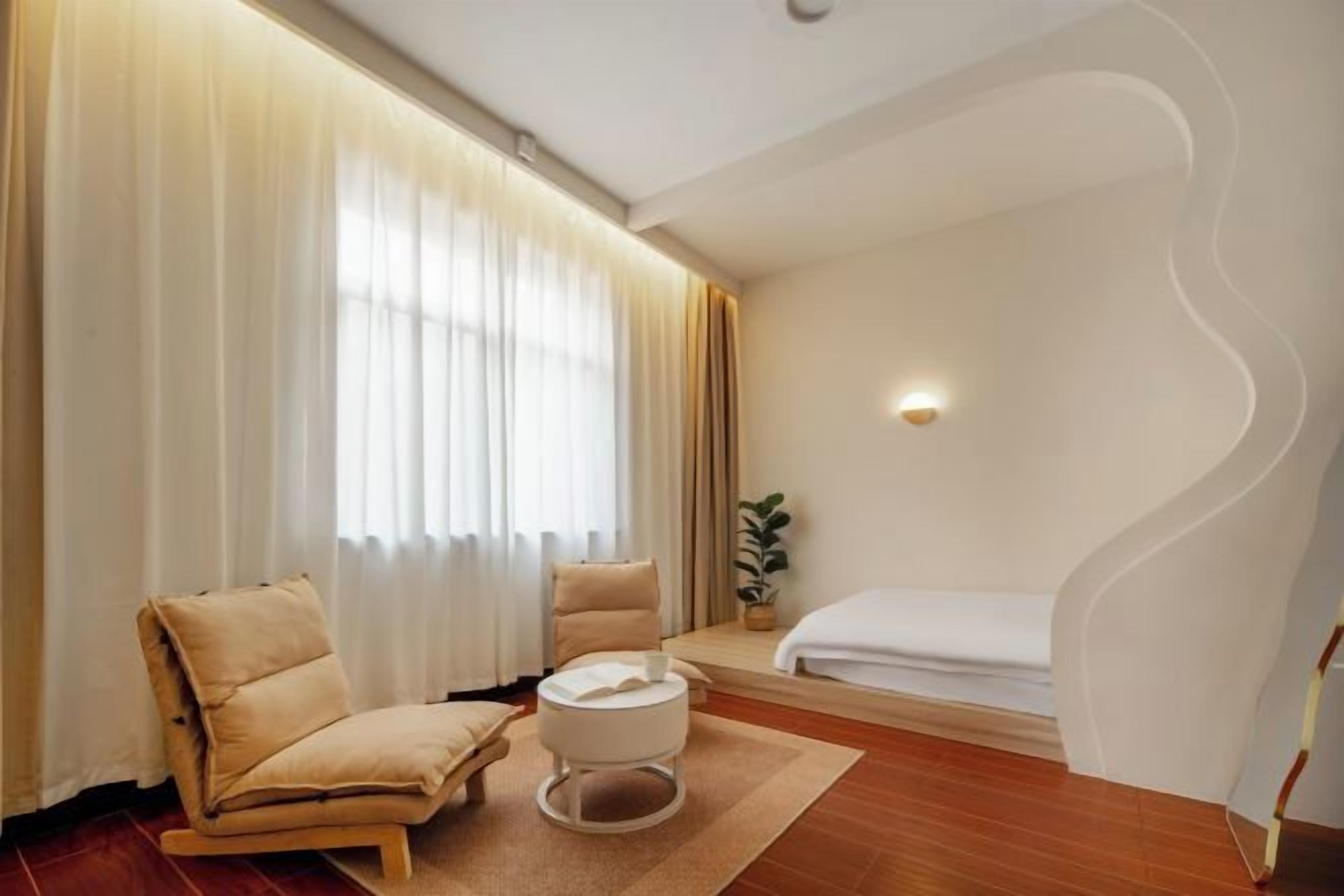 A hotel room with a bed, two chairs, a small table, a potted plant, and a wall-mounted light.