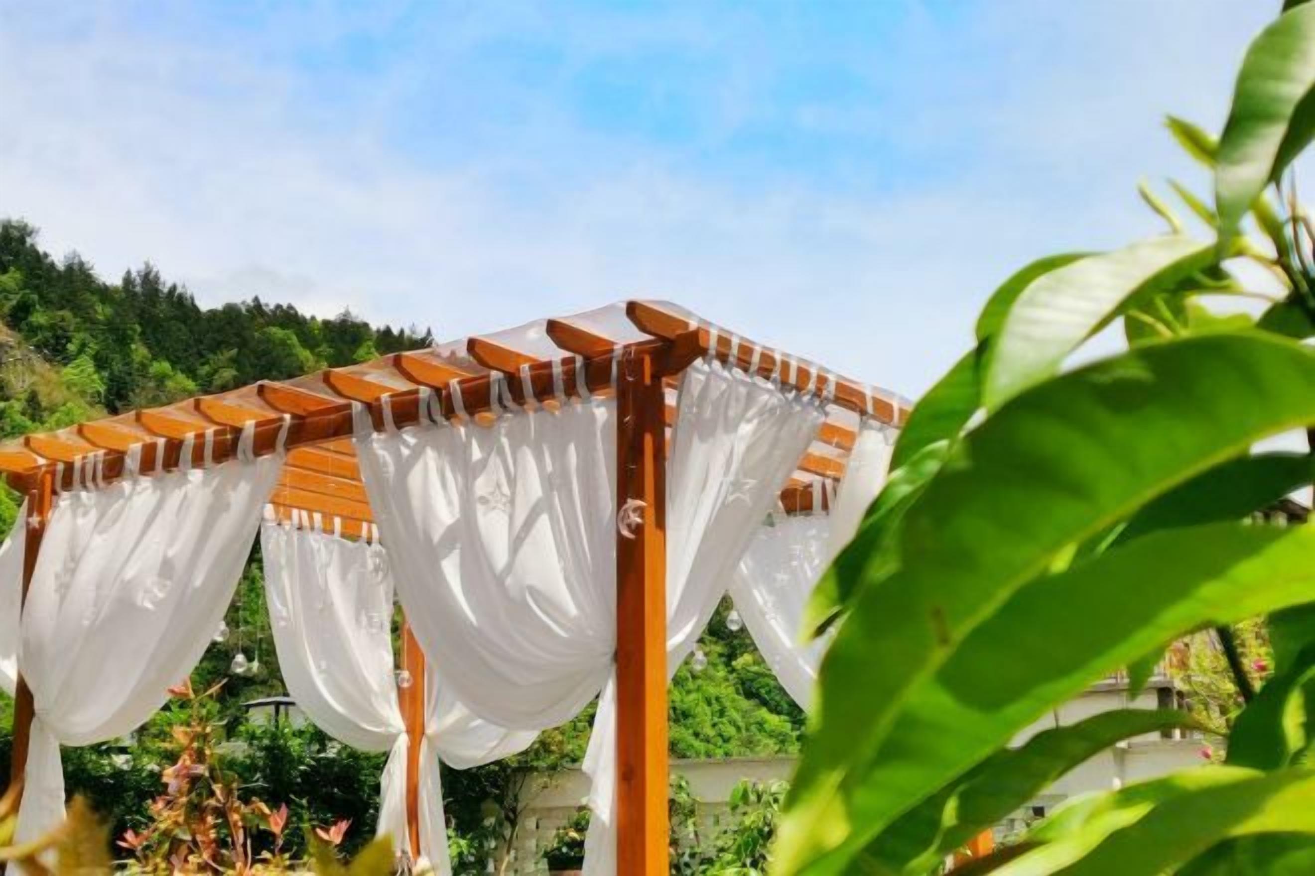 A wooden pergola with white drapes, set against a backdrop of greenery and a clear sky.