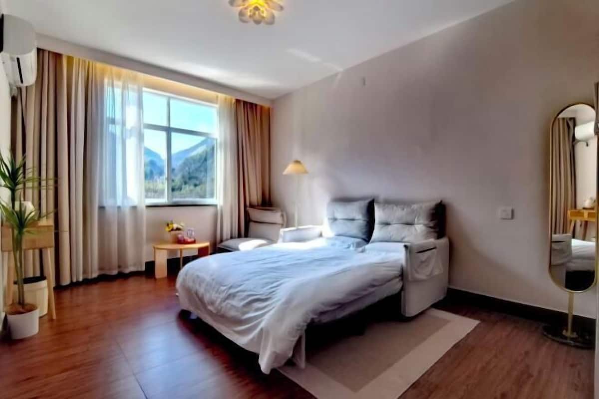 A modern hotel room with a large bed, a bedside table, a lamp, a mirror, and a window with curtains.