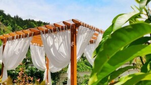 Una pérgola de madera con cortinas blancas, rodeada de vegetación.