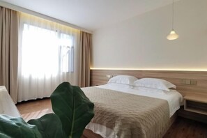 Habitación de hotel con cama, mesita de noche, lámpara colgante y una planta verde grande.