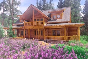 Une cabane en rondins dotée d’un balcon, d’une véranda et d’un jardin où poussent des fleurs violettes.