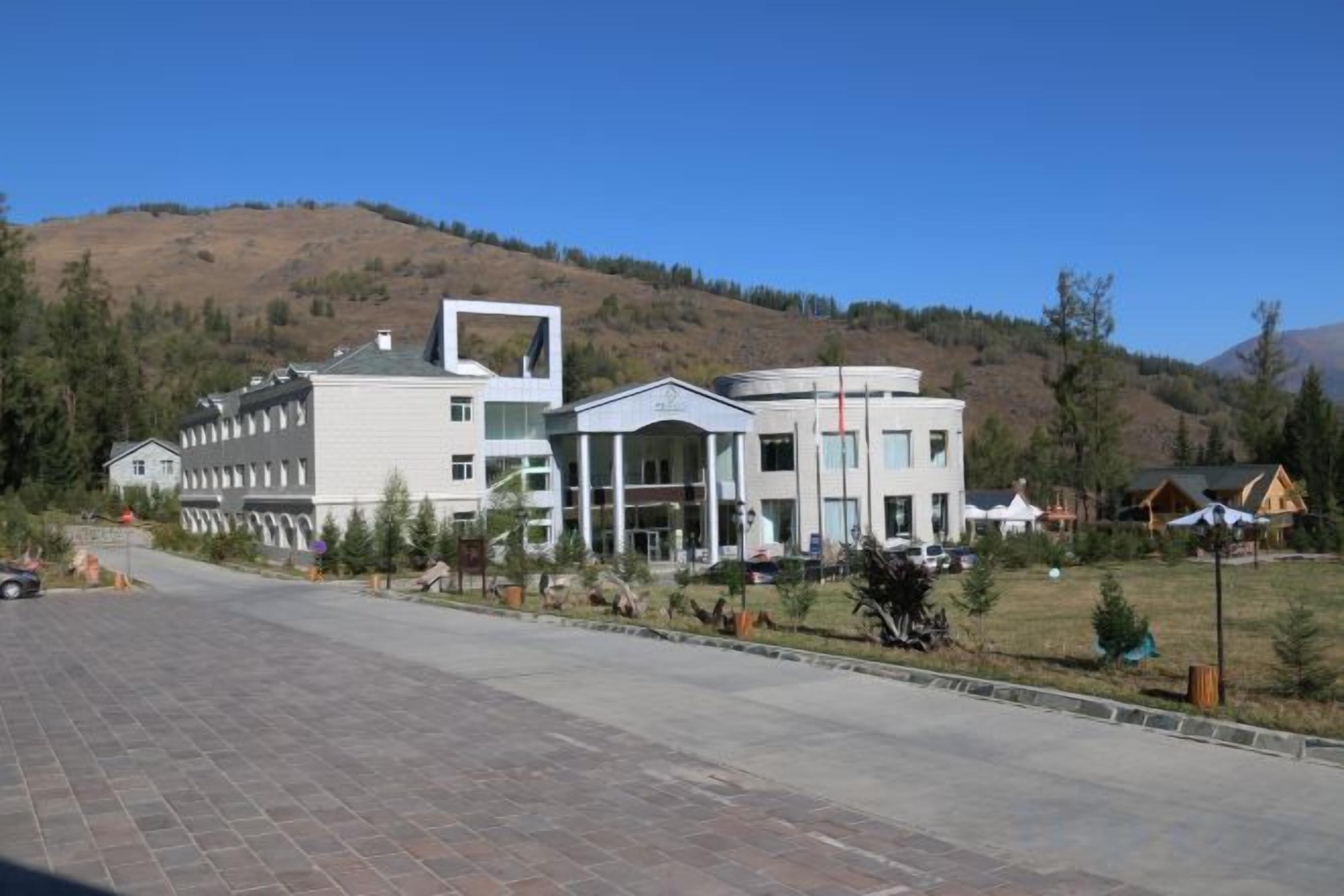 A modern building with a central atrium, surrounded by a paved area and a hilly landscape.