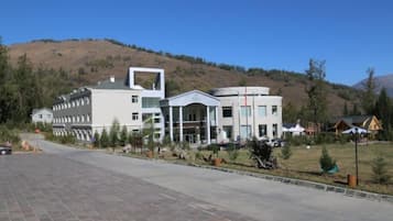 A modern building with a central atrium, surrounded by a paved area and a hilly landscape.