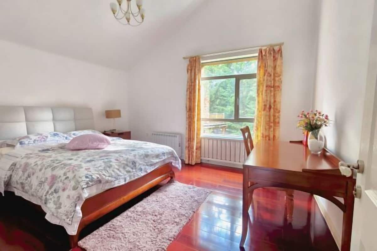 Un dormitorio con cama, una mesa de madera, una silla, una ventana con cortinas y un radiador.