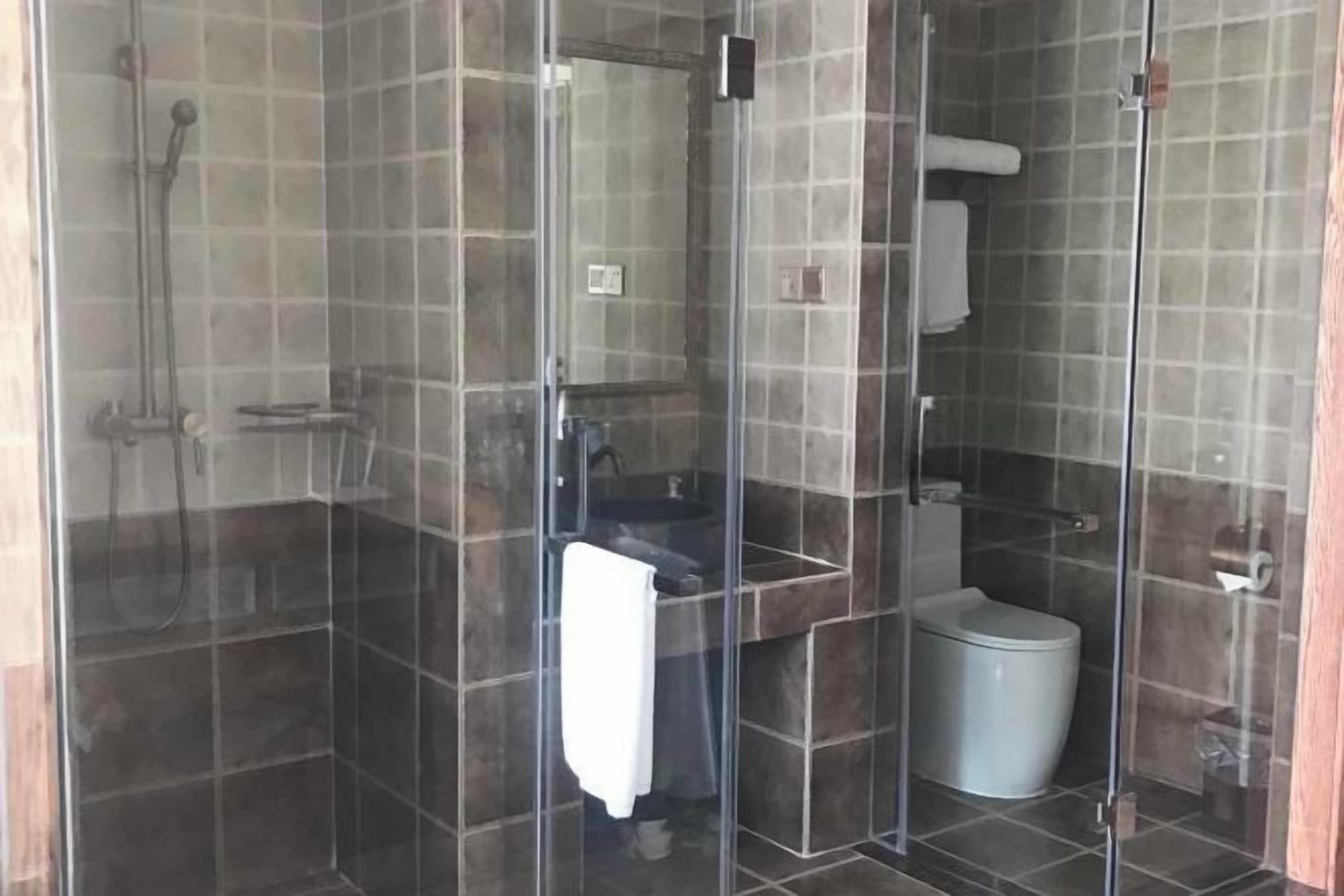 Área de chuveiro com paredes de vidro, piso ladrilhado, pia e vaso sanitário.