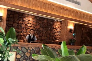 Mostrador de recepción con pared de piedra de fondo, una recepcionista y plantas en macetas.