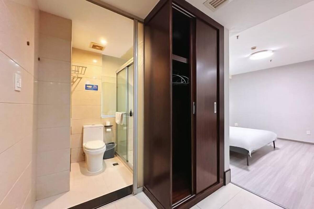 A modern hotel room with a glass shower enclosure, a white toilet, a bed, and a wooden wardrobe.