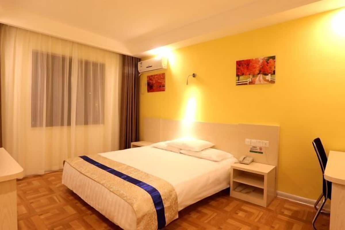A hotel room with a bed, bedside table, lamp, and a wall-mounted air conditioner.