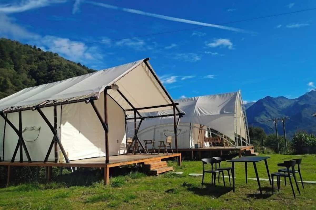 Varias tiendas de lona con bancos y mesas de madera, ubicadas en un área cubierta de césped con montañas al fondo.
