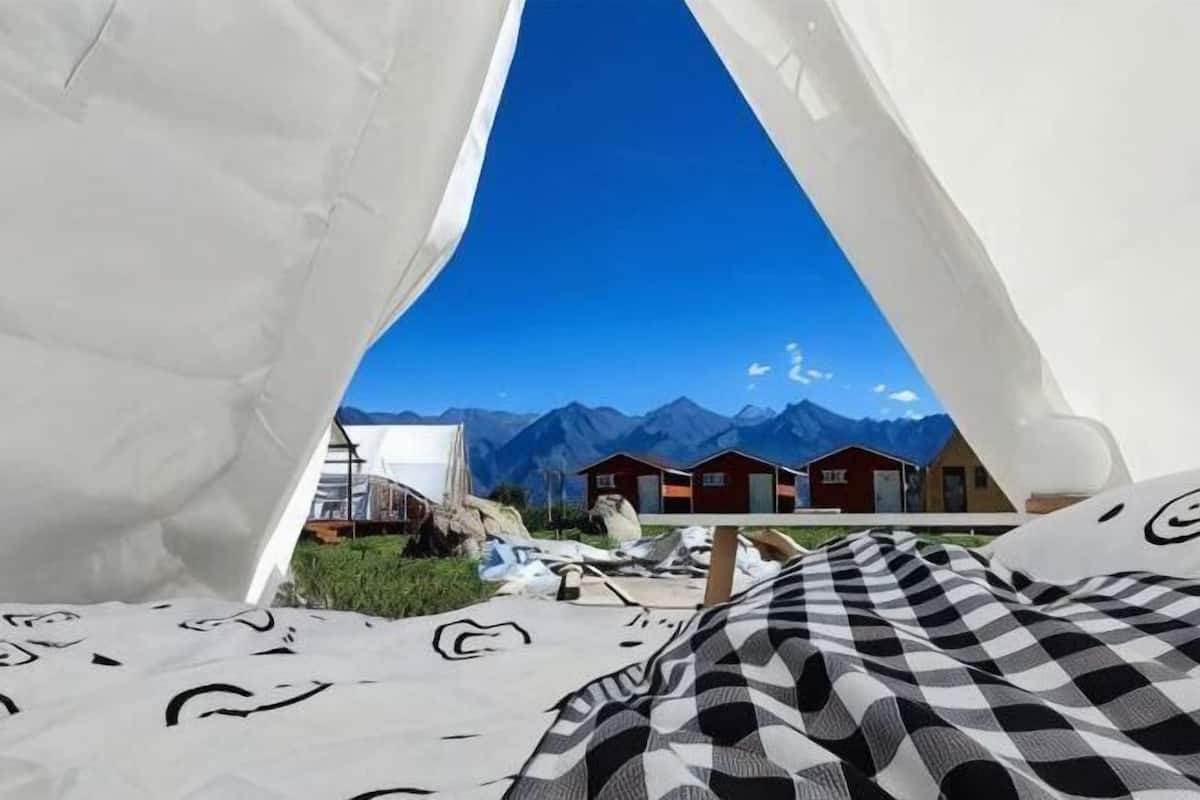 Una tienda de campaña con vista a la montaña, una cama con manta a cuadros y una estructura de madera pequeña al fondo.