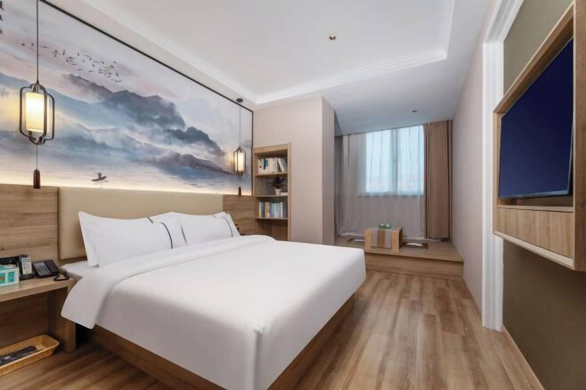 A modern hotel room with a large bed, a desk with a telephone, a bookshelf, and a painting of a landscape.